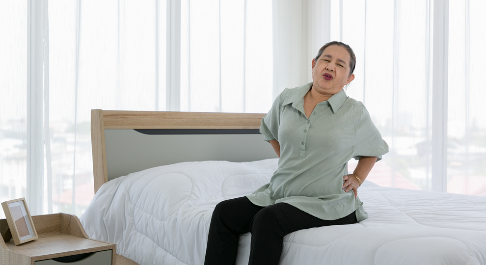 Older woman in a light green blouse sits on a white bed, leaning back with hands on hips in a bright bedroom with sheer curtains and a wooden nightstand nearby.