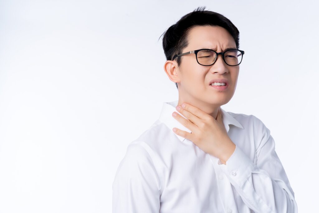 Young man in a white shirt wearing glasses grimaces and touches his throat, showing discomfort against a pale background.